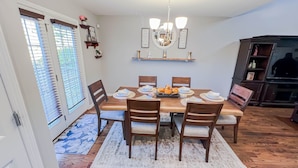 A dining room with a wooden table set for six, surrounded by chairs, a chandelier, and wall-mounted shelves with decorative items.