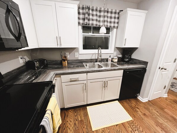A kitchen with white cabinets, a black countertop, a window with a checkered valance, and a wooden floor.