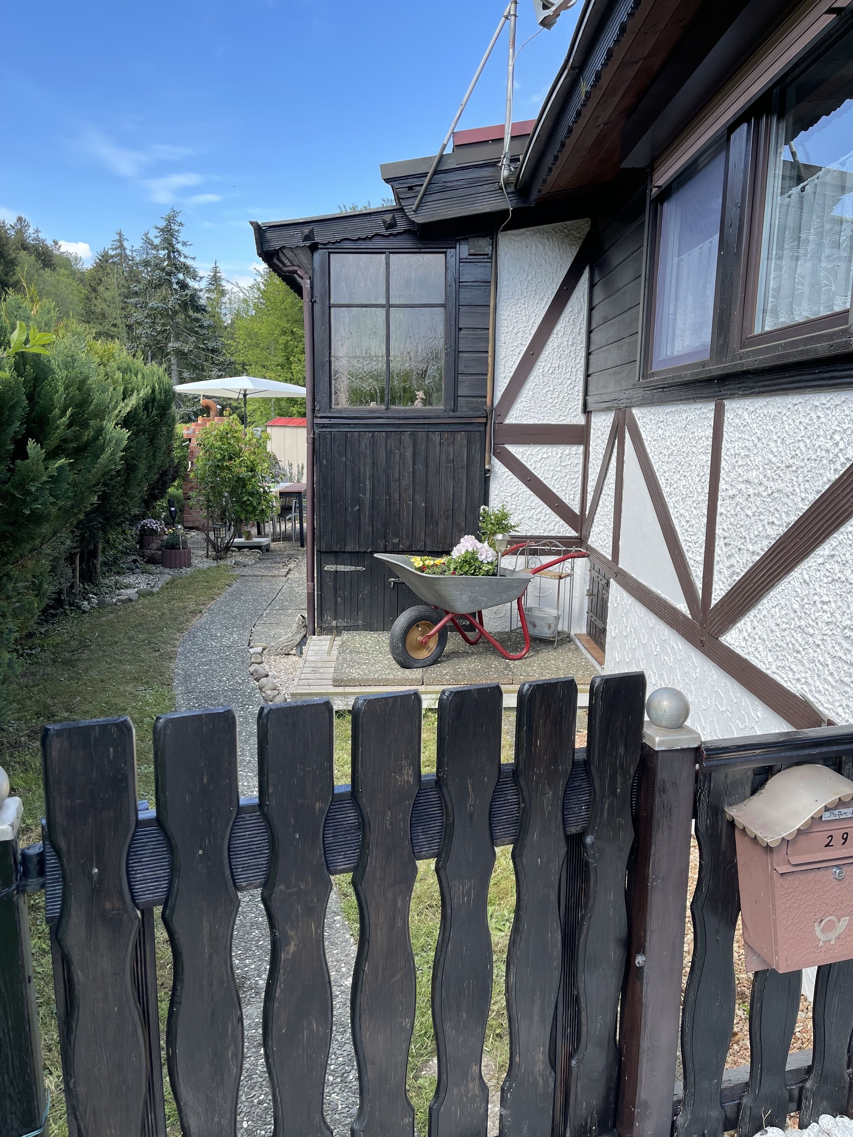 Ein Haus mit Holzfassade, einer Schubkarre voller Pflanzen und einem umzäunten Garten.