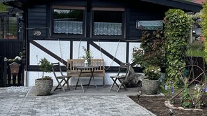 Ein rustikales Holzgebäude mit schwarzem Dach und weißem Zierprofil, im Vordergrund eine Bank und Kübelpflanzen.