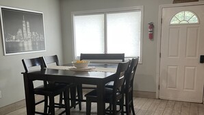 Zona pranzo con tavolo e sedie in legno, una foto incorniciata di un panorama urbano sulla parete, un'ampia finestra con persiane e una porta bianca con decorazione.