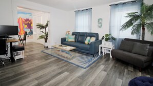 Een woonkamer met een blauwe bank, een bruine bank, een salontafel en een televisie. Er staan twee potplanten en er hangt een schilderij aan de muur.