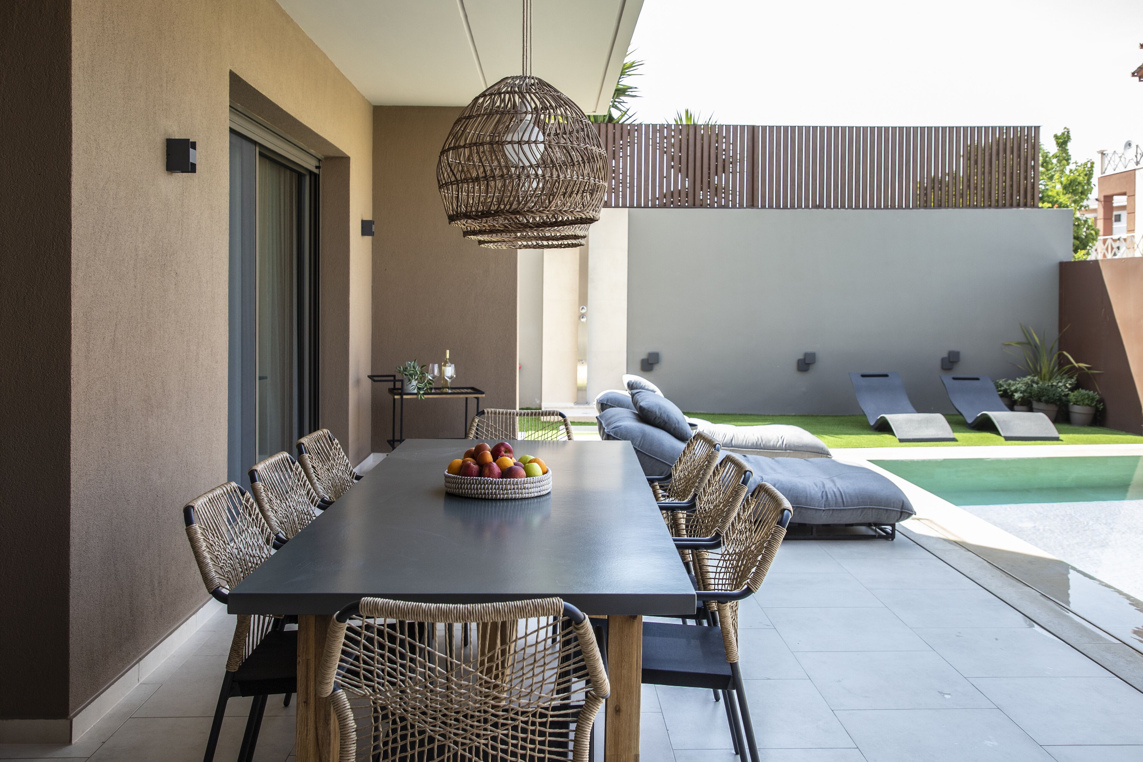 A modern outdoor dining area with a long table, wicker chairs, and a fruit bowl. There is a pool and lounge chairs in the background.