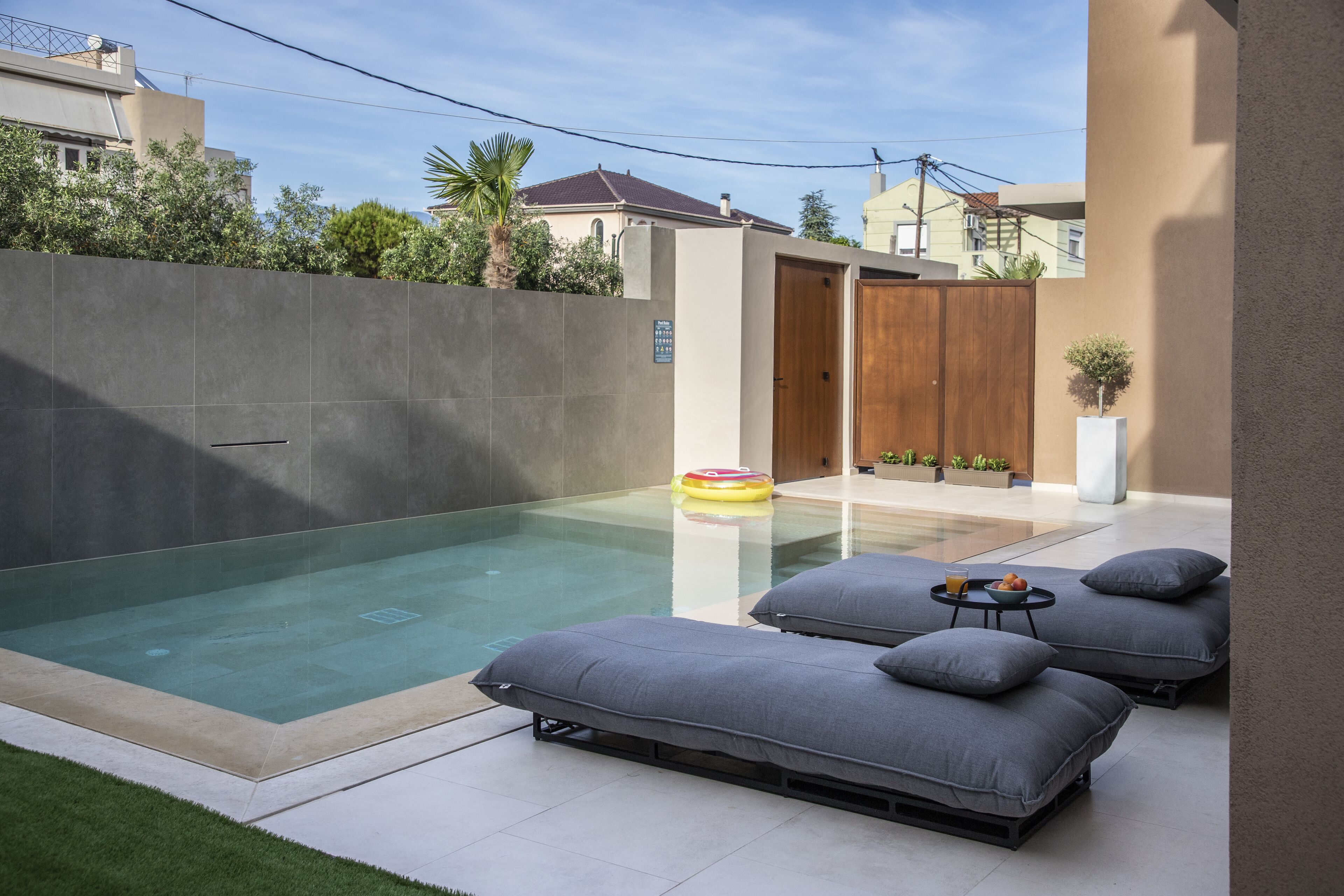 Zona de piscina moderna al aire libre con cerramiento de vidrio, dos sillones de descanso grises y una mesa pequeña con una bebida.