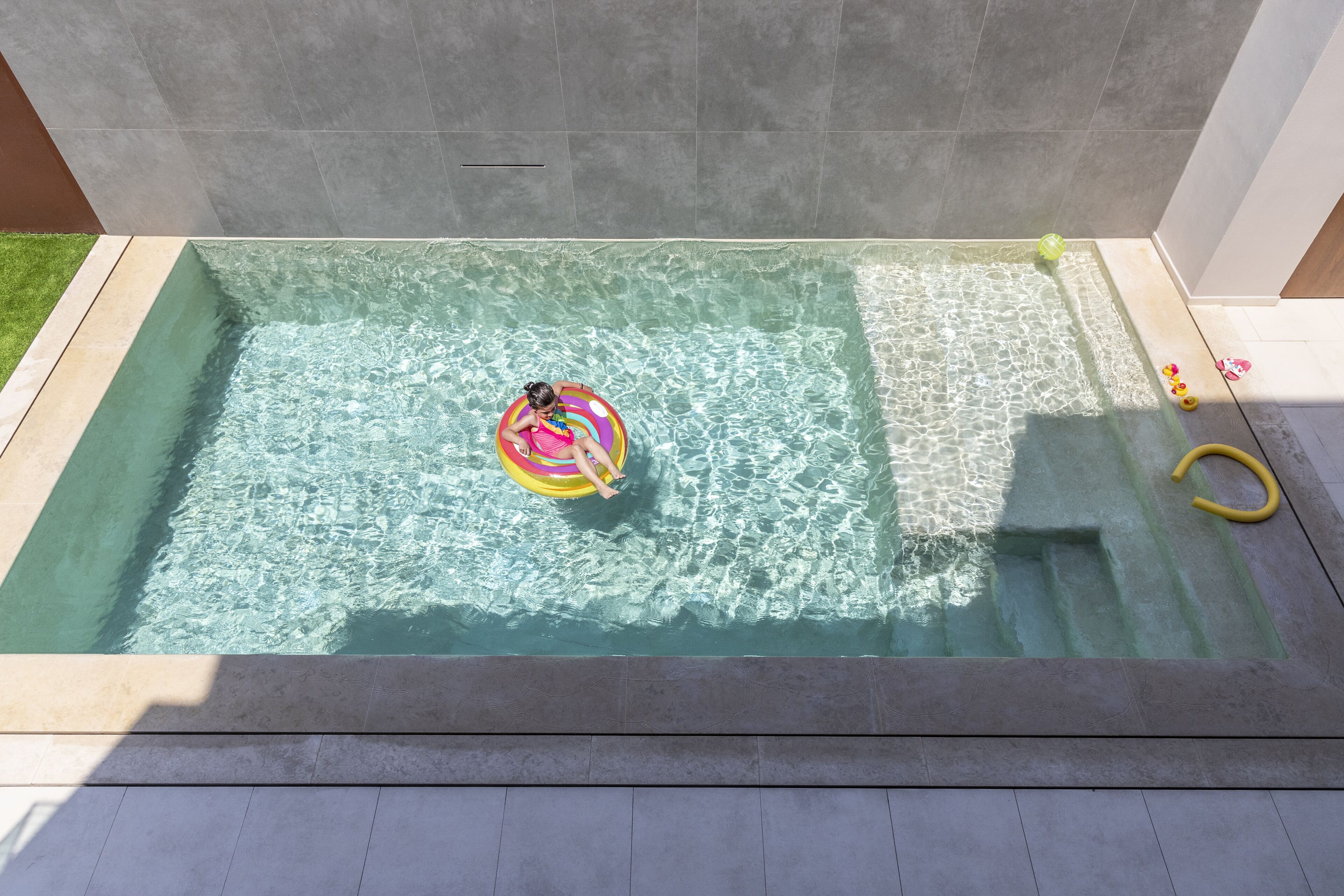 A rectangular hot tub with clear water, a person on a colorful float, and a yellow hose on the right side.