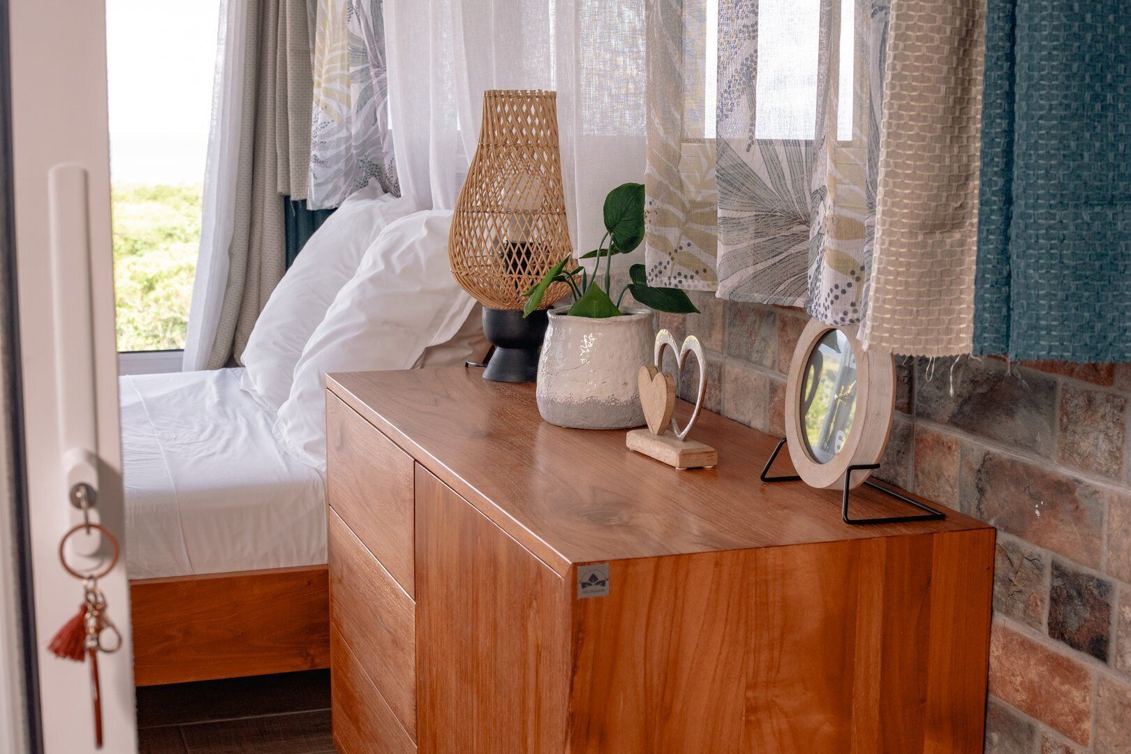 A bedroom with a wooden dresser, a wicker lamp, a potted plant, a decorative heart-shaped object, and a mirror on the dresser.
