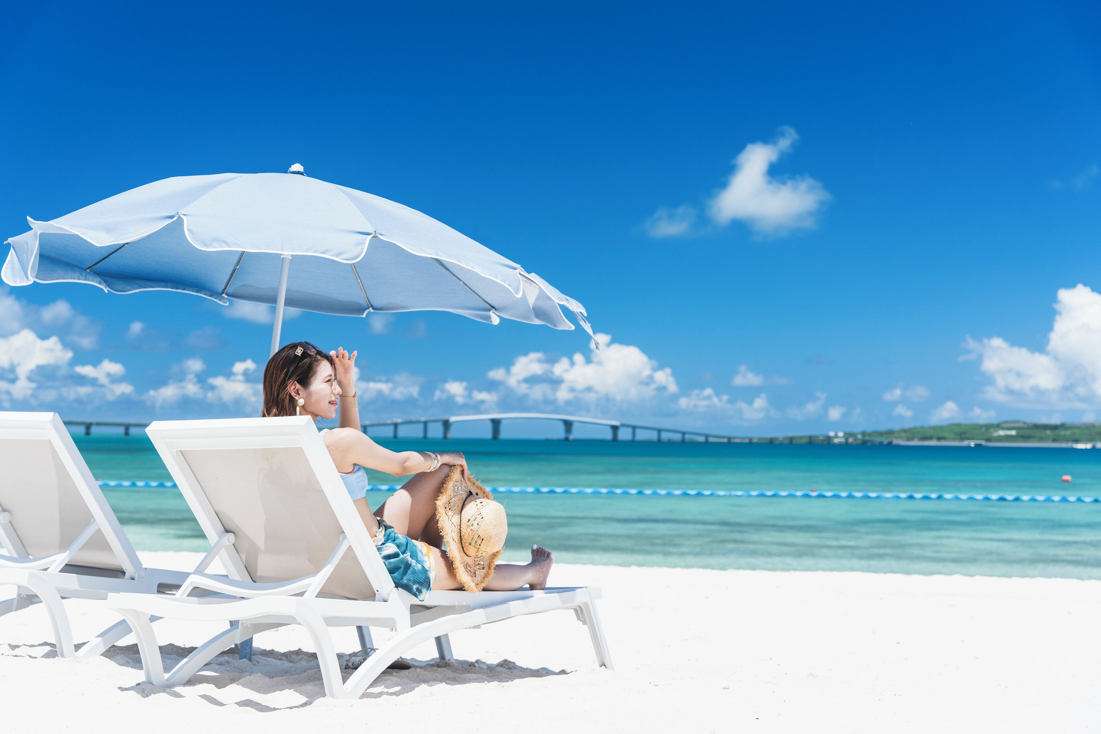 A beach scene with a person sitting under a white umbrella, white lounge chairs, and a clear blue sky.