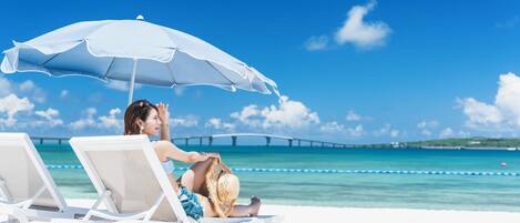 A beach scene with a person sitting under a white umbrella, white lounge chairs, and a clear blue sky.