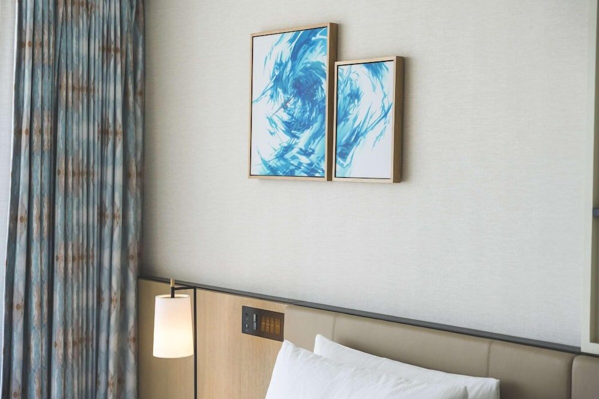 A hotel room with a bed, a headboard, a wall-mounted light, and two framed artworks.