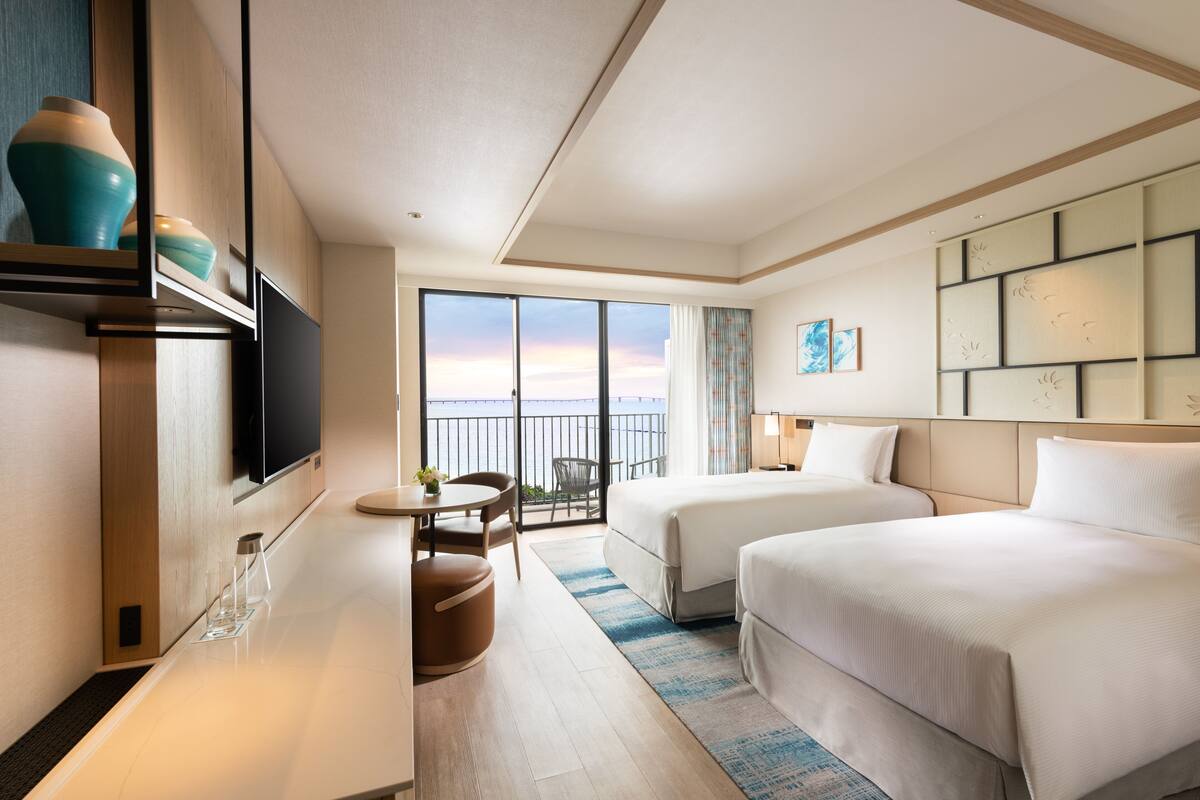 Habitación de hotel con dos camas, televisor, zona de comedor y balcón con vistas.