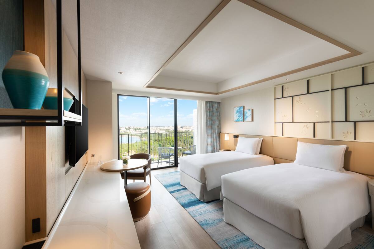 Habitación de hotel con dos camas, un escritorio, una silla, un televisor y un ventanal con vistas.
