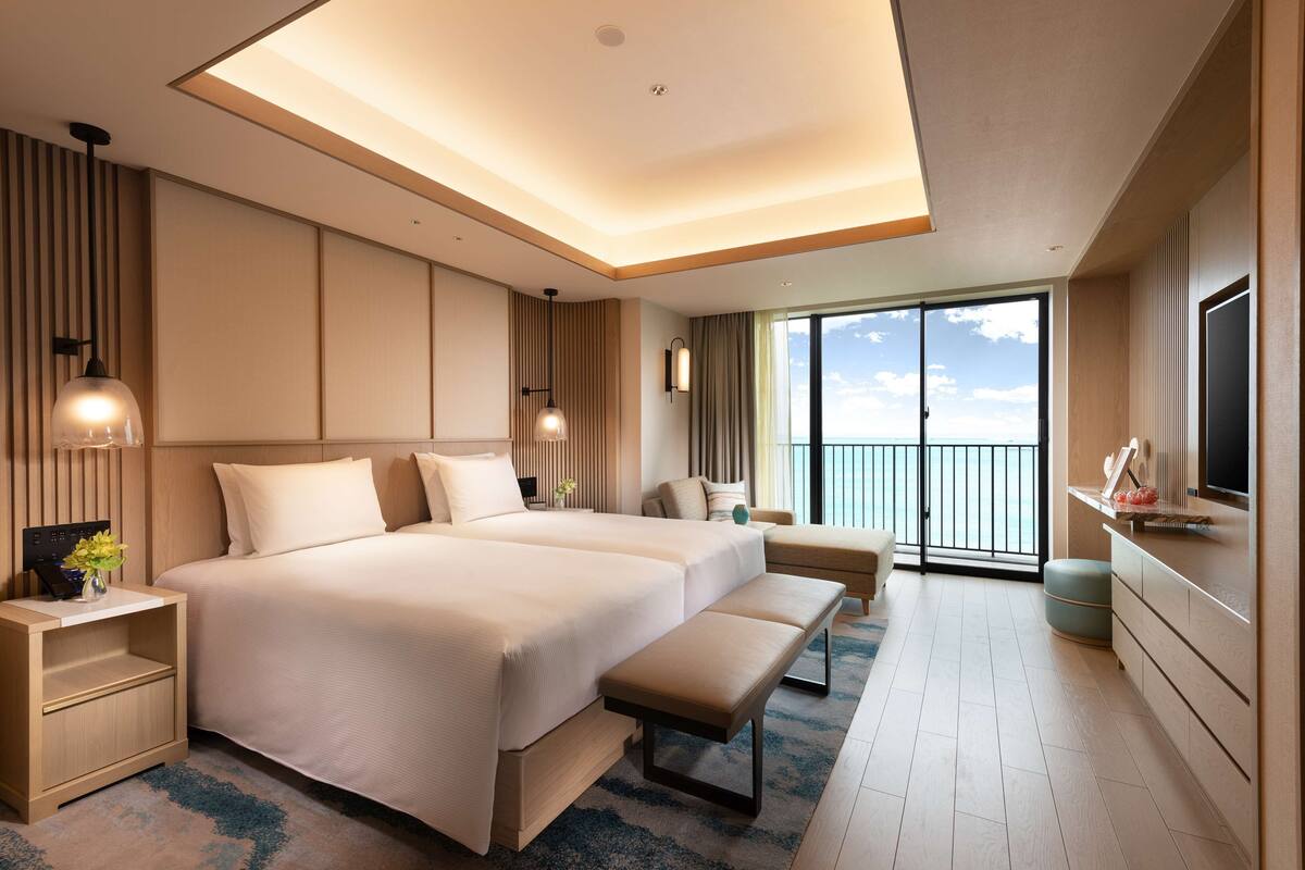 Una habitación de hotel moderna con una cama grande, mesitas de noche, un banco, un televisor y un balcón con vistas.