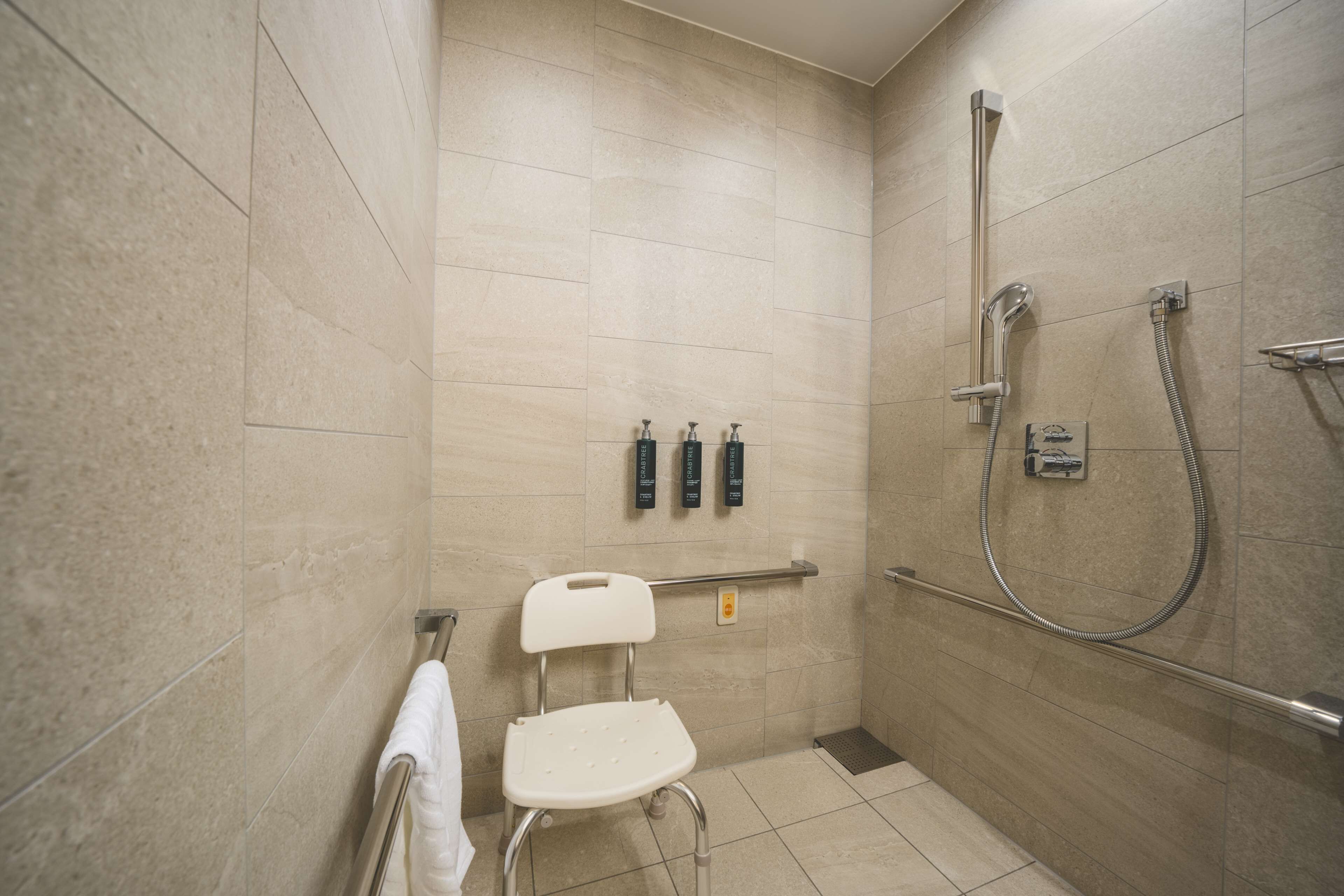 A small, modern bathroom with beige tiles, a white chair, a towel rack, and a handheld showerhead.