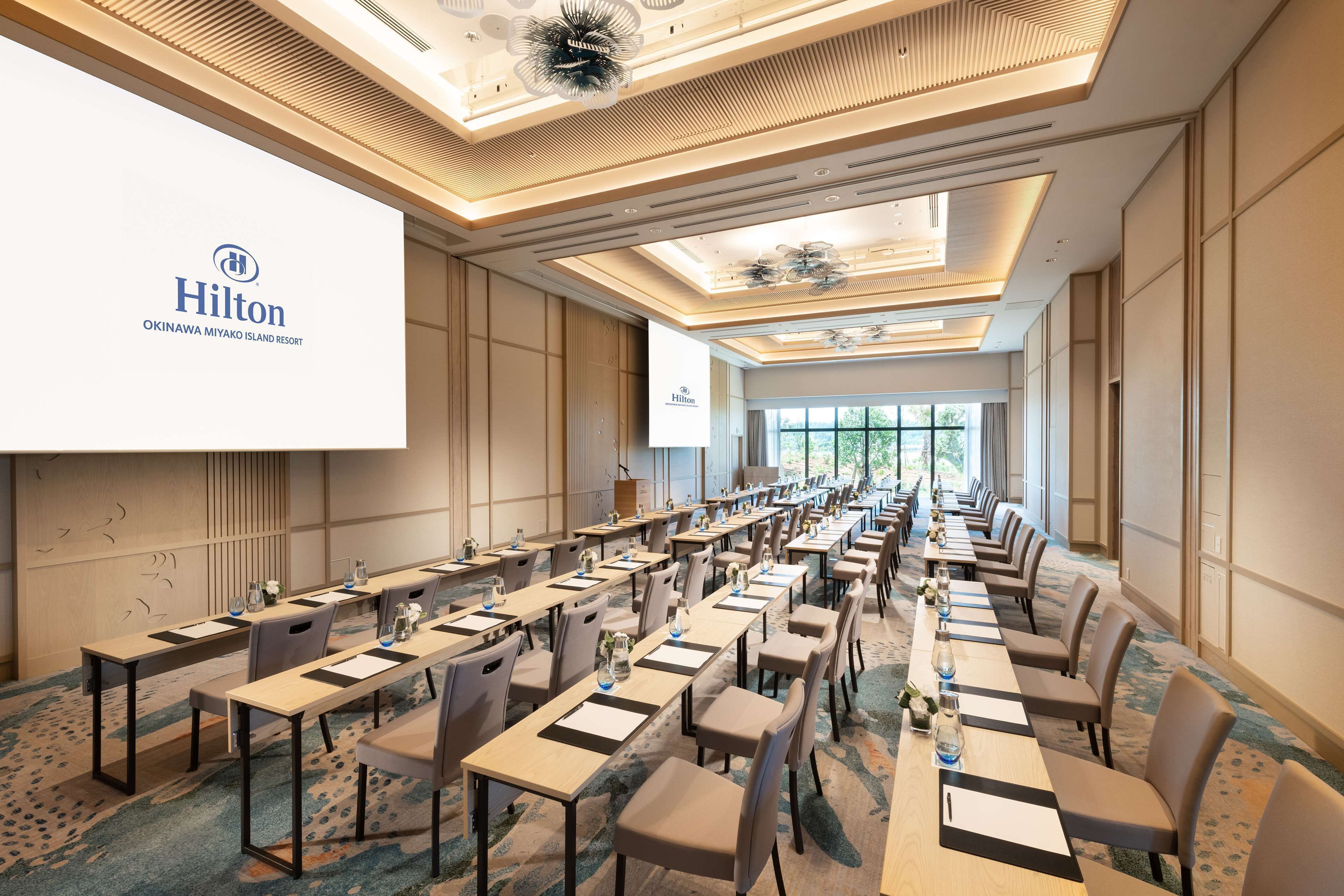 Een congrescentrum in het Hilton Okinawa Miyako Island Resort met lange tafels, stoelen en een groot scherm waarop de naam van het hotel staat.