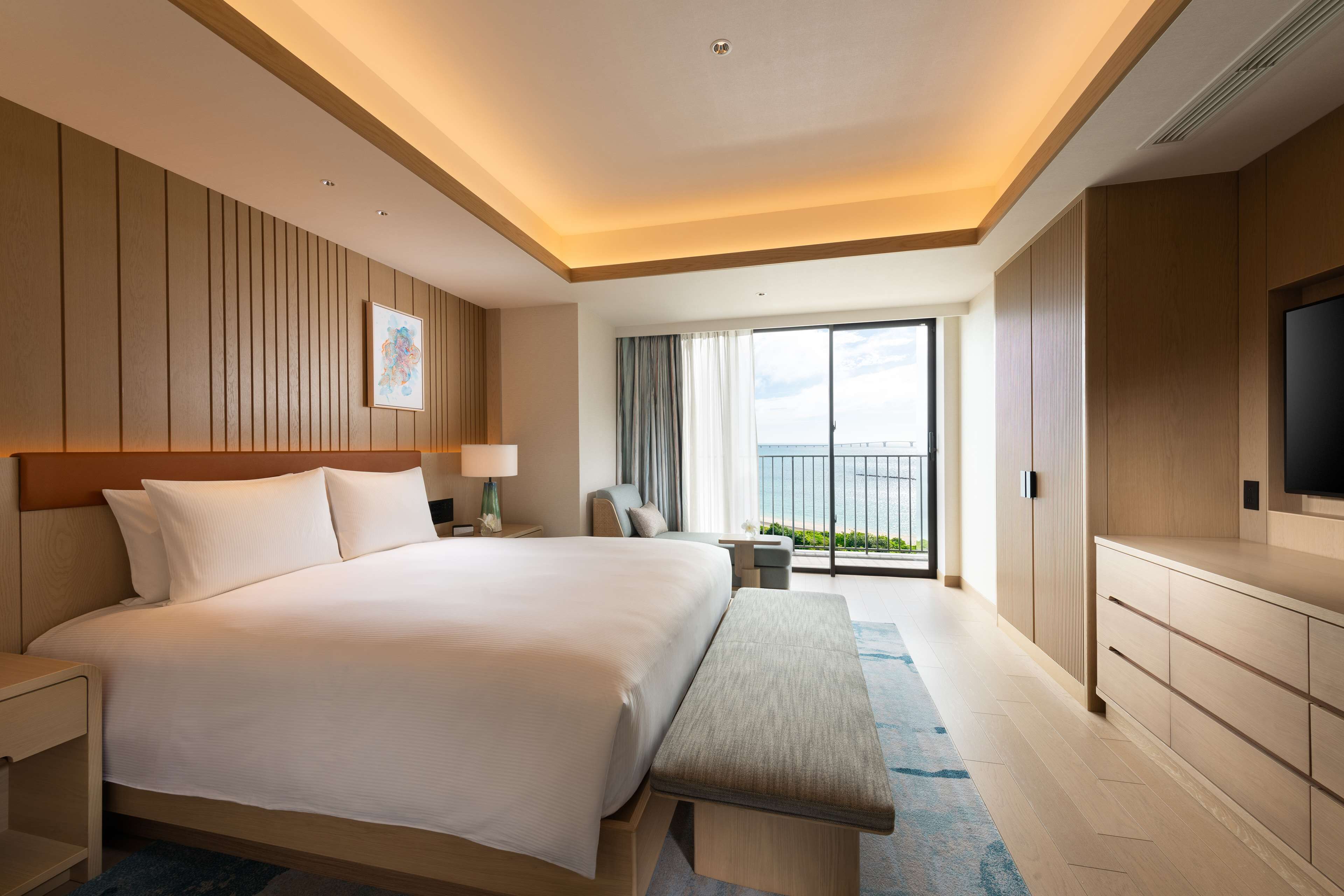 A modern hotel room with a large bed, a bench, a TV, and a balcony with a view.