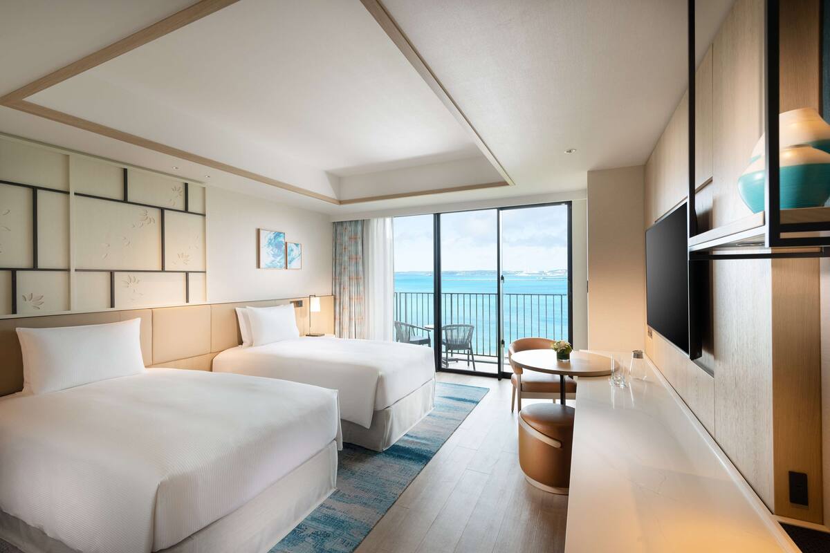 Habitación de hotel con dos camas, zona de comedor, televisión y balcón con vistas al mar.