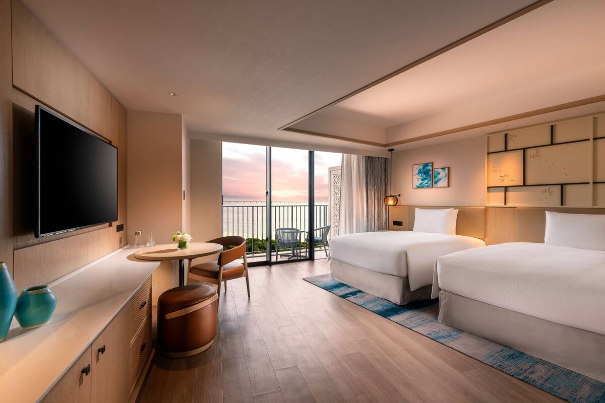 Habitación de hotel con televisor, una mesa de comedor, dos camas, balcón con vistas y una obra de arte grande en la pared.