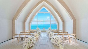 An aisle with chairs and floral arrangements, leading to a large arched window with a cross, overlooking a body of water. - Hilton Okinawa Miyako Island Resort (Miyakojima)