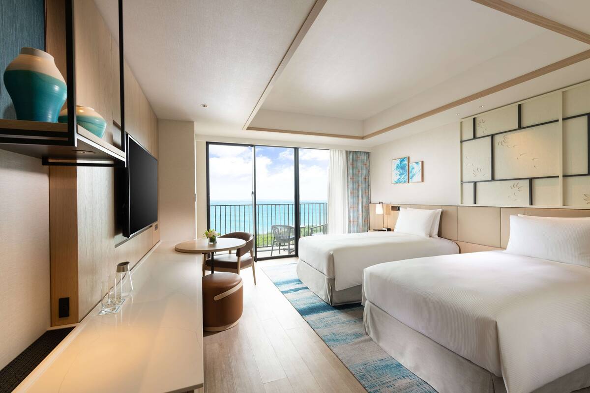 Habitación de hotel con dos camas, televisión, una pequeña mesa con sillas y un balcón con vista al mar.