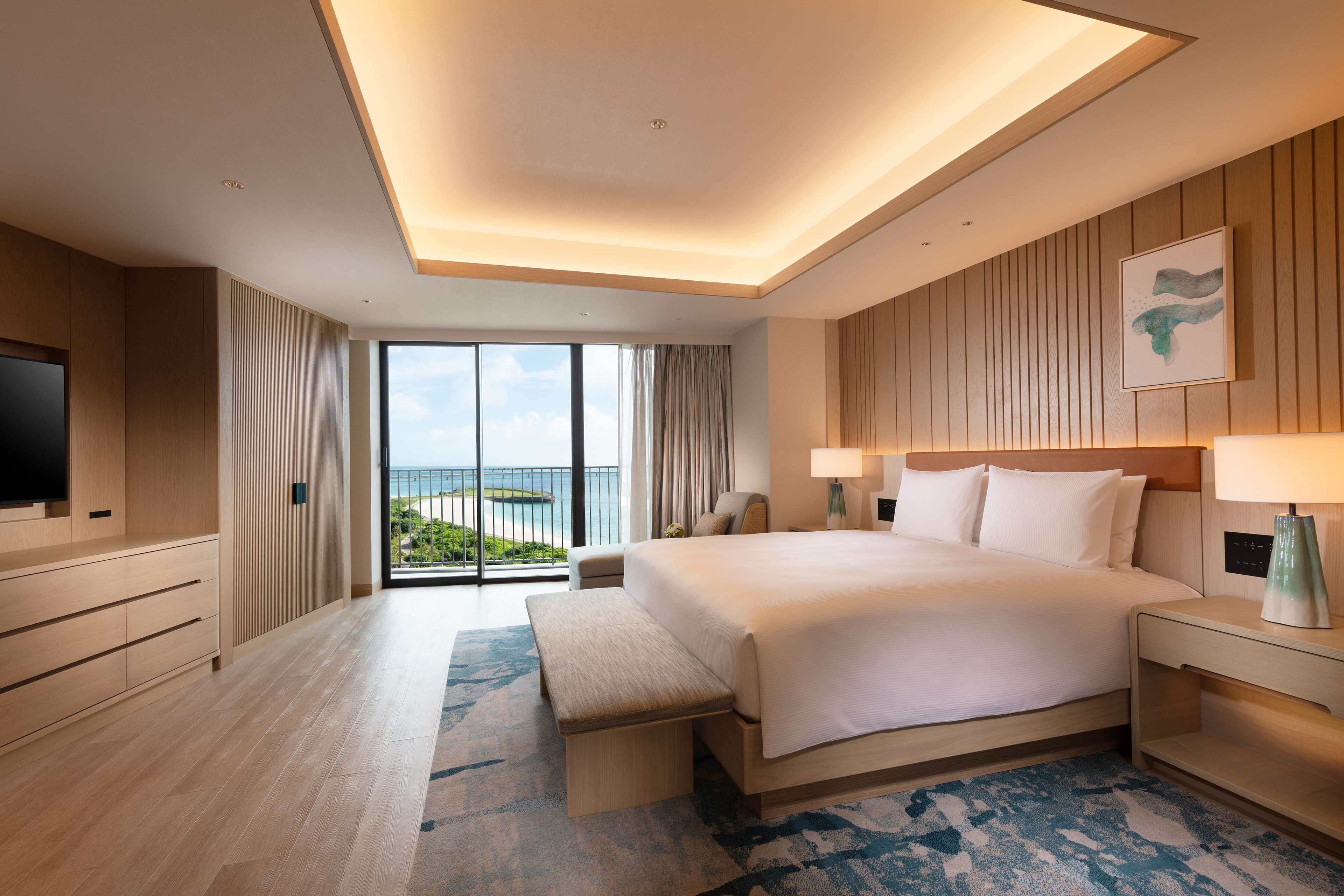 Una habitación de hotel moderna con una cama grande, un televisor de pantalla plana, una banca y vista al océano.