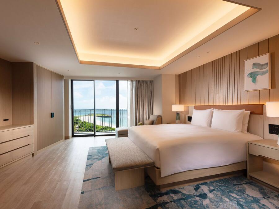 A modern hotel room with a large bed, a flat-screen TV, a bench, and a view of the ocean.