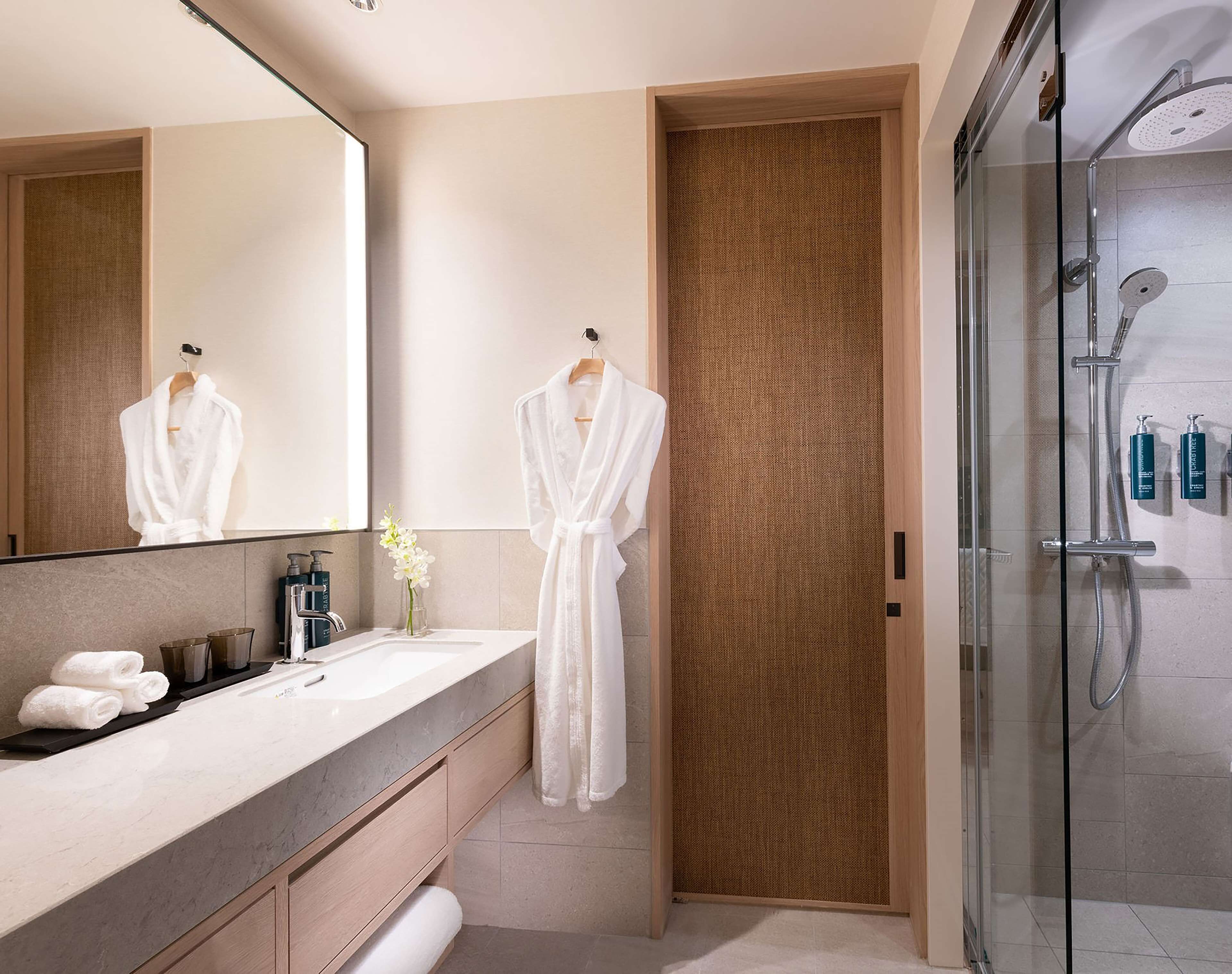 A modern bathroom with a large mirror, two white bathrobes hanging, a glass shower enclosure, and a marble sink.
