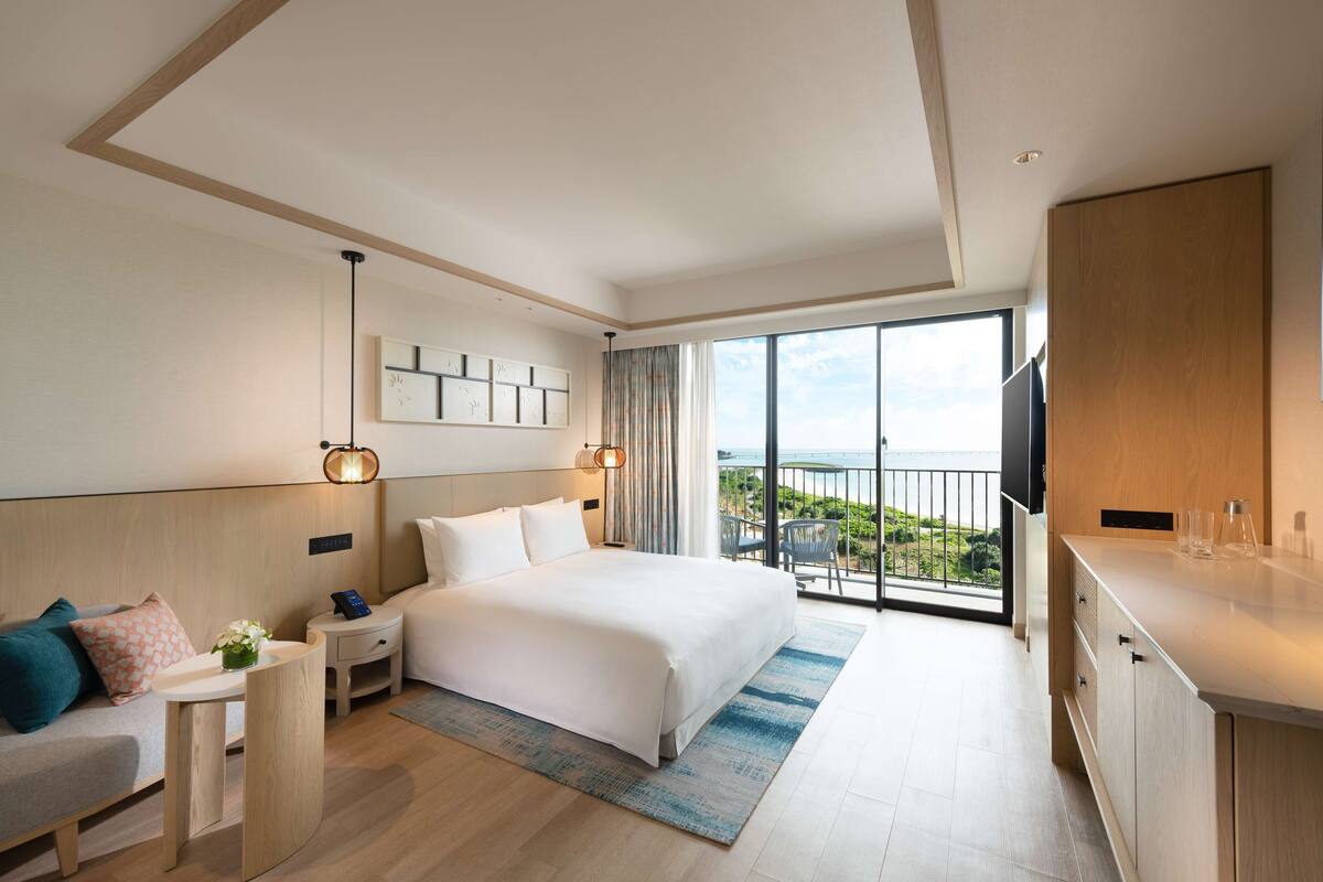 Habitación de hotel moderna con una cama grande, un escritorio, una silla, un televisor y un balcón con vistas.