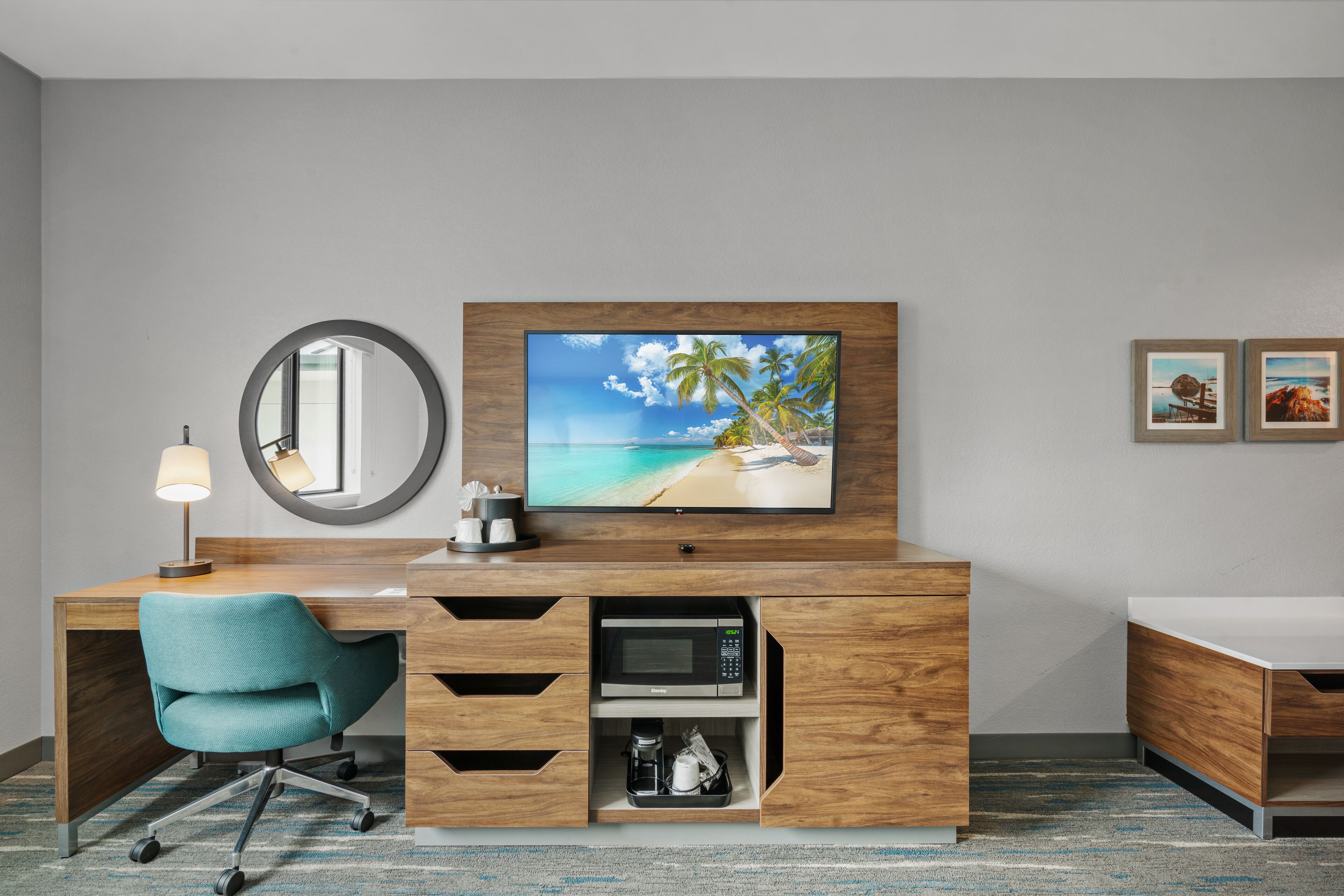 Ein Hotelzimmer mit einem Holztisch, einem blaugrünen Bürostuhl, einem Flachbildfernseher, einer Mikrowelle und einer Kaffeemaschine.