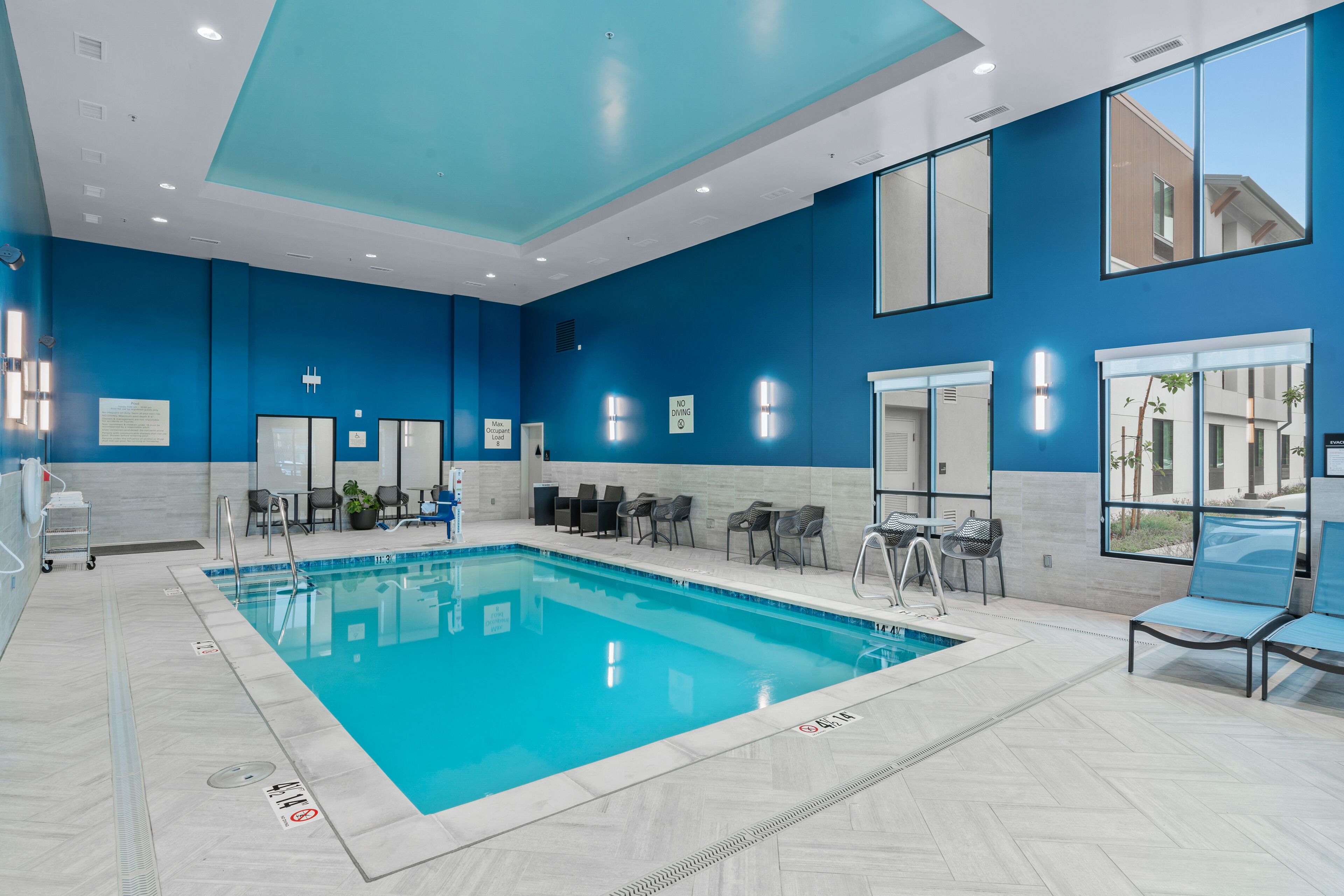 Una piscina interior moderna con paredes azules y agua cristalina, rodeada de zonas de estar y un mostrador de recepción.