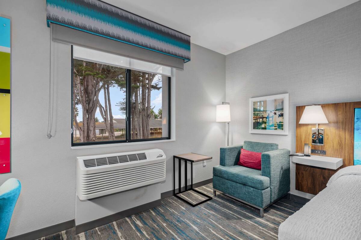 A hotel room with a large window, a standing heater, a blue armchair with a red pillow, a bedside table, and a lamp.