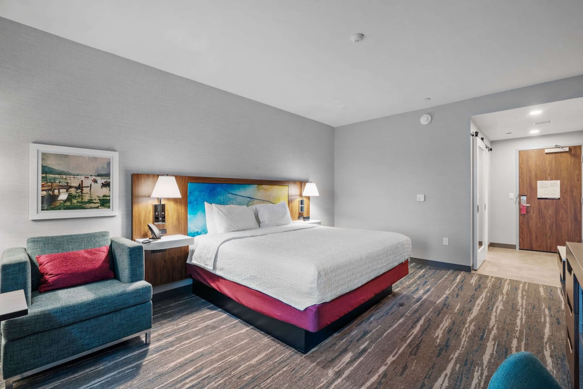 A modern hotel room with a large bed, a desk, a chair, and a painting on the wall.