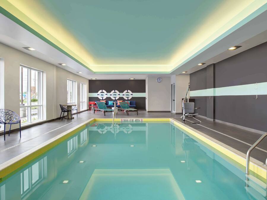 Piscina interior con agua cristalina, zona poco profunda señalizada y área de descanso con sillas y mesas.