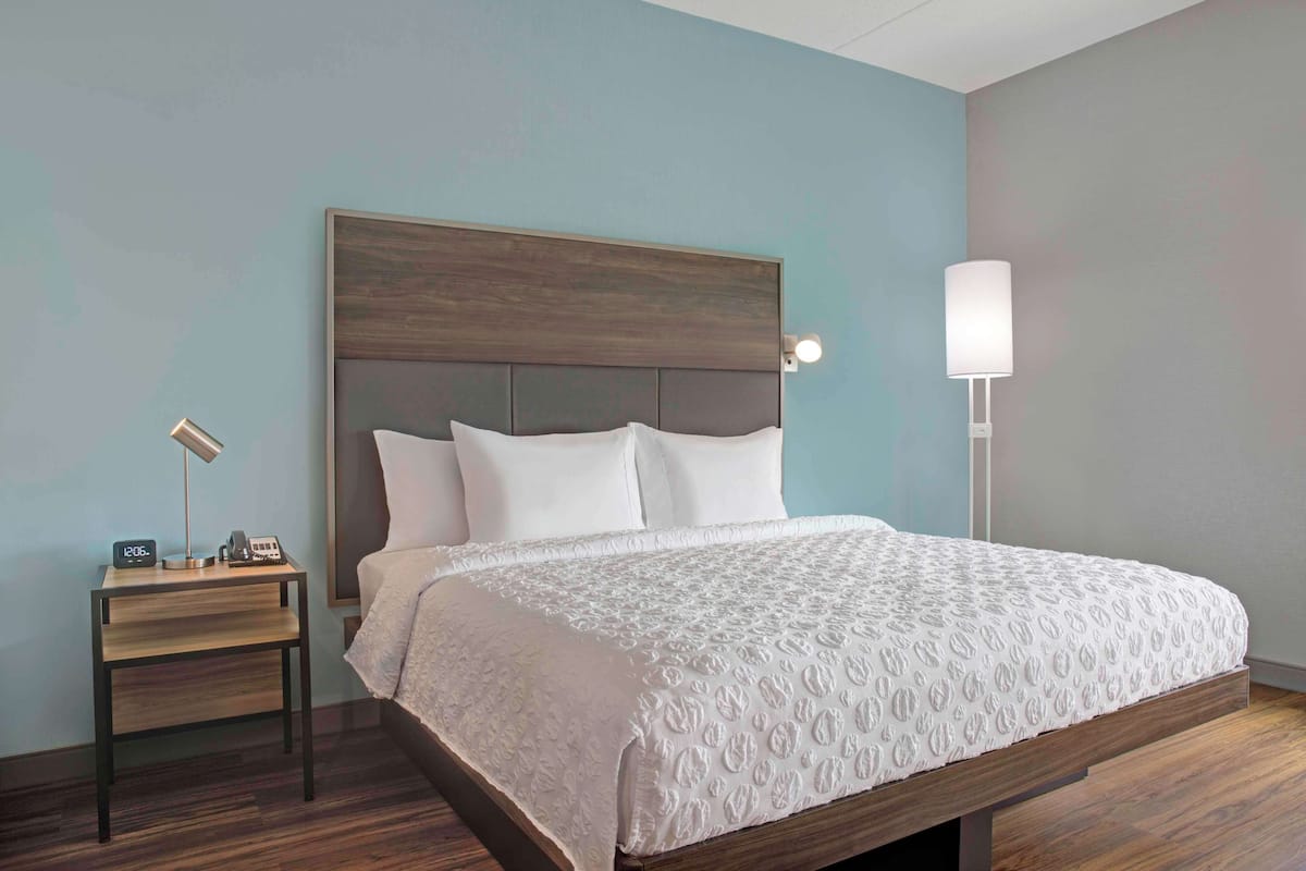 Een net opgemaakt bed met een houten hoofdbord, een nachtkastje met een lamp en een wandlamp.