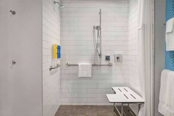 A small, white-tiled shower room with a handheld showerhead, a towel rack, and a small table.