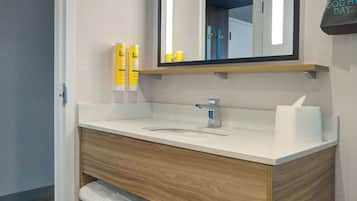 A modern bathroom with a wooden vanity, a sink, and a mirror. There are toiletries on the vanity and a towel rack below.