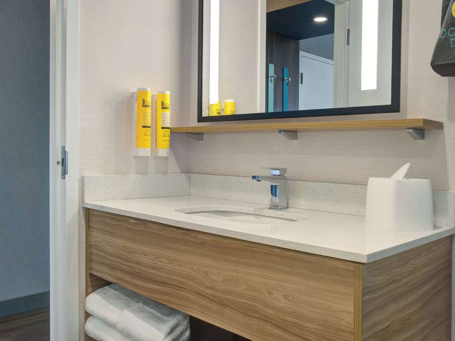A modern bathroom with a wooden vanity, a sink, and a mirror. There are toiletries on the vanity and a towel rack below.