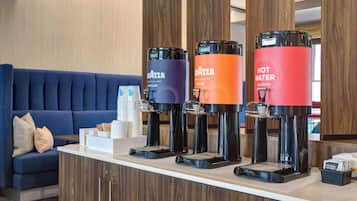 Zona de recepción de un hotel con un sofá azul, una estación de café con cuatro dispensadores etiquetados 'IvaZZA' y un dispensador de 'Agua Caliente'.