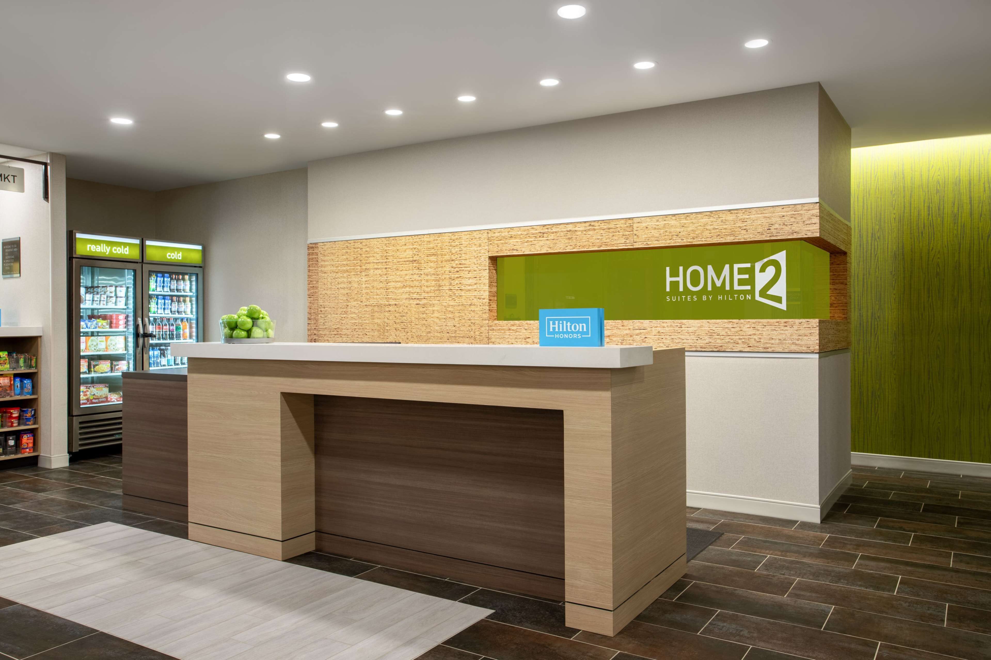 En hotellreception med en skylt som visar texten ”HOME 2 SUITES BY HILTON” och en ”Hilton Honors”-märke. Det finns ett kylskåp med drycker och ett fat med frukt.