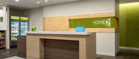 En hotellreception med en skylt som visar texten ”HOME 2 SUITES BY HILTON” och en ”Hilton Honors”-märke. Det finns ett kylskåp med drycker och ett fat med frukt.