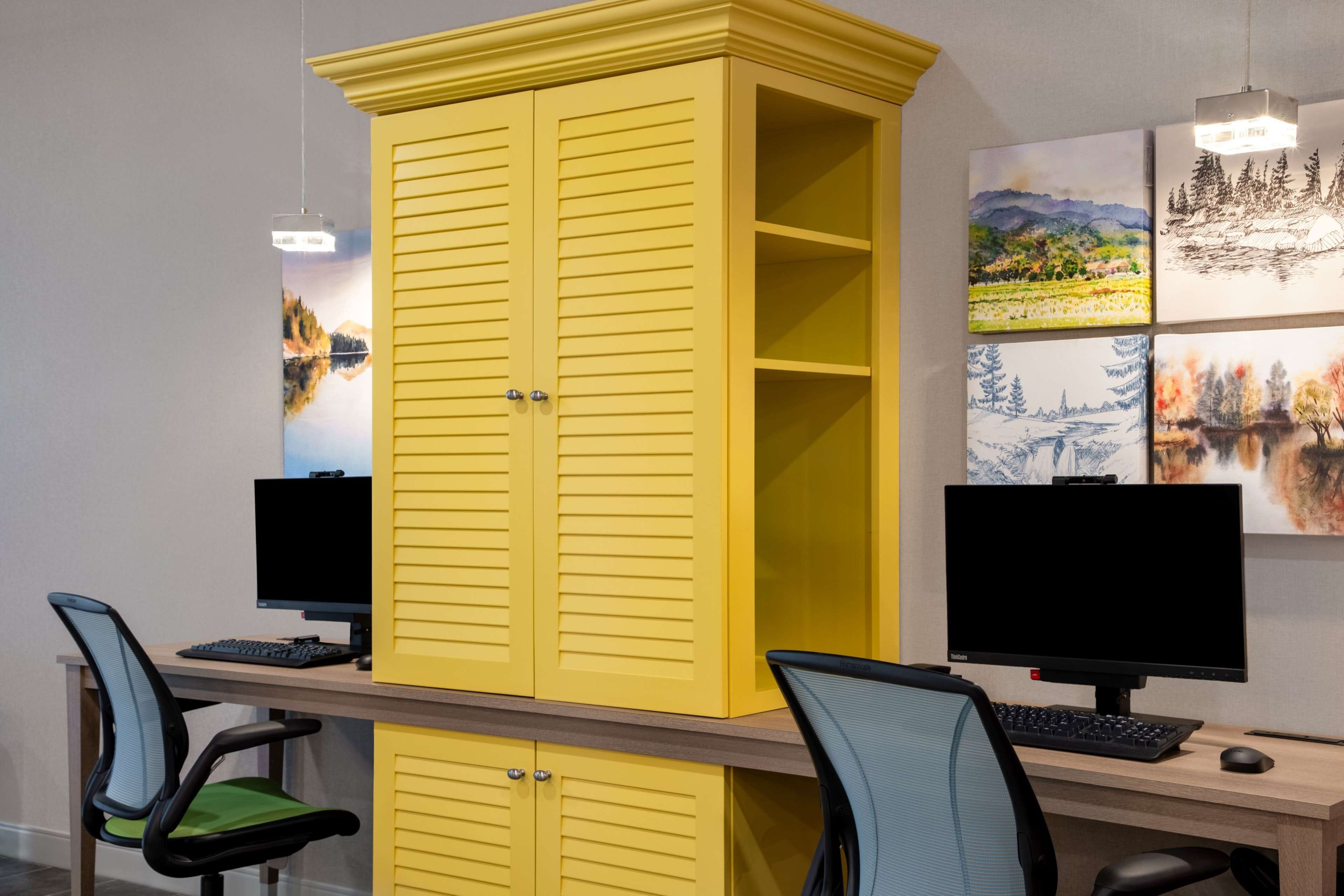 Un espace de travail comprenant deux bureaux, chacun équipé d’un écran d’ordinateur, d’un clavier et d’une souris. On y trouve deux chaises, dont une avec un coussin vert. Un meuble jaune à deux portes se trouve à l’arrière-plan.
