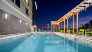 A swimming pool with blue tiles, surrounded by lounge chairs and umbrellas, and a building with a sign in the background. - Home2 Suites By Hilton Calhoun (Calhoun)