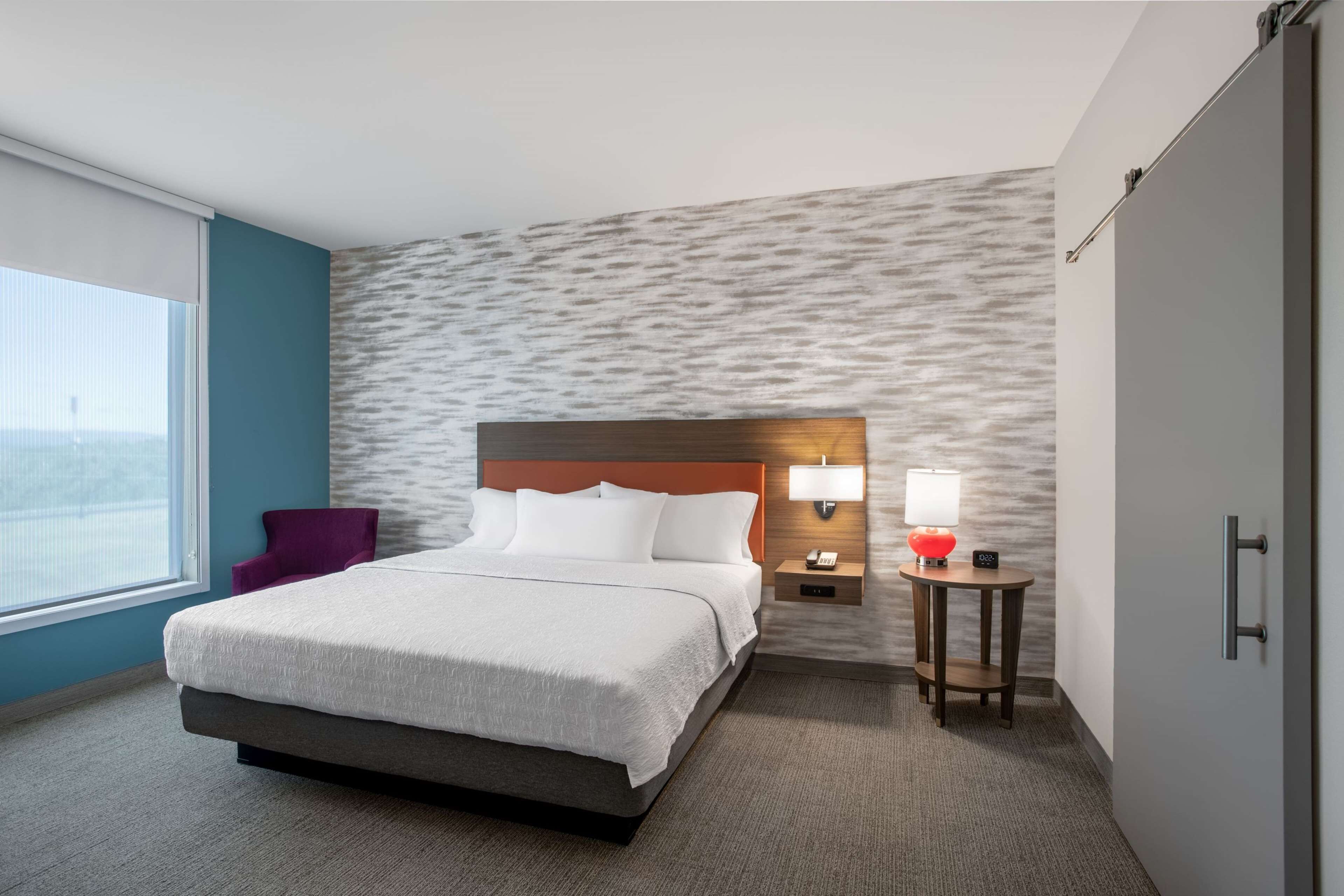 A hotel room with a bed, a nightstand with a lamp, a chair, and a wardrobe.
