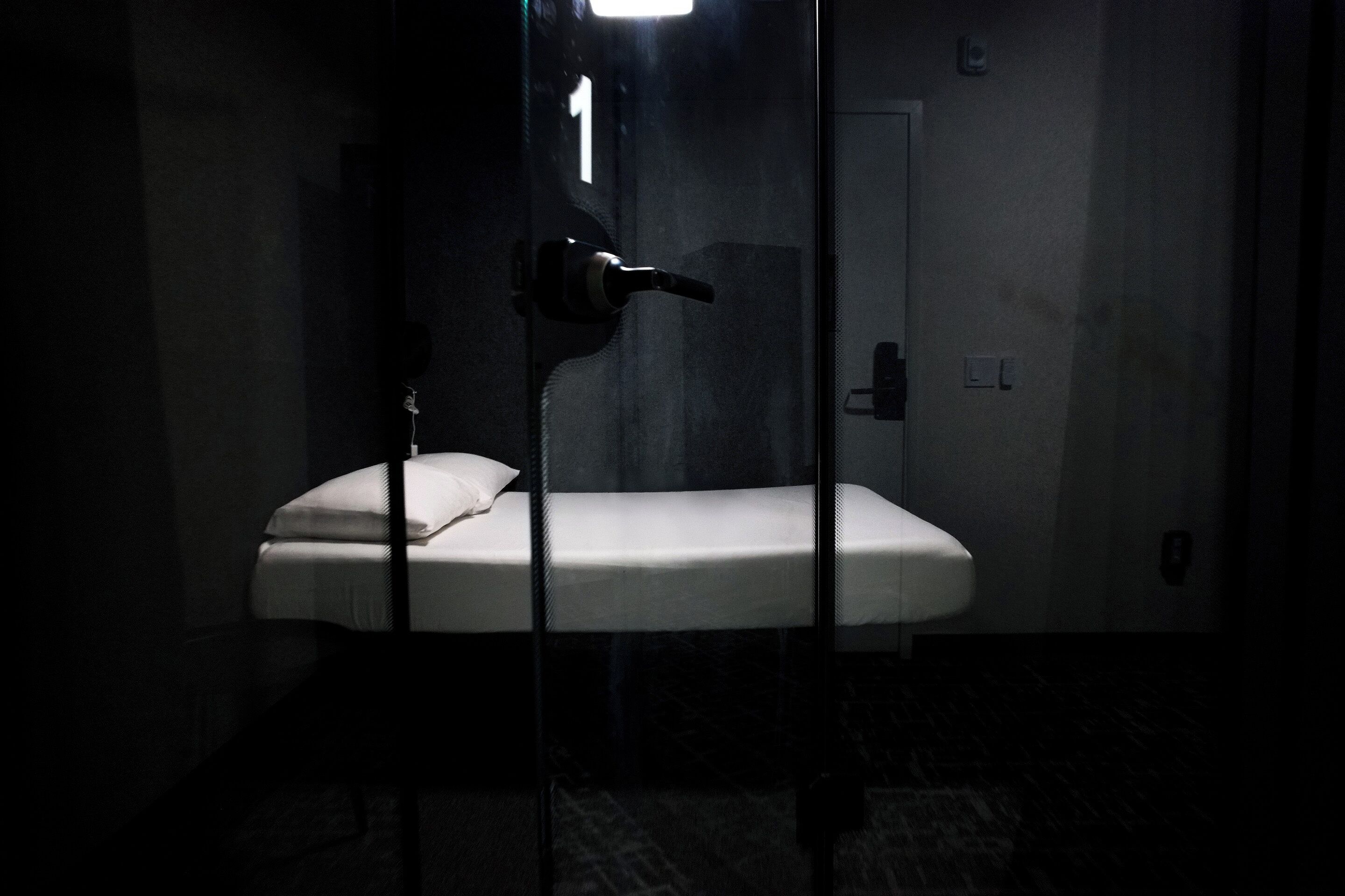 A dimly lit room with a single bed, a visible door, and a wall-mounted light fixture.
