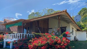 A wooden house with a thatched roof, a porch with a white railing, and vibrant red flowers in the foreground. - Relaxed house in Well located, Authentic, tranquil and unspoilt fishing village. (Tonosi)