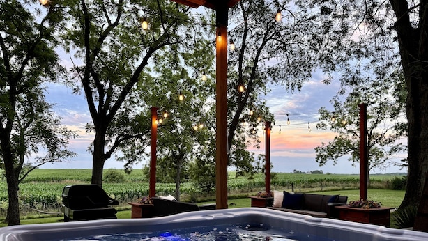 Un jacuzzi con vista a un campo y árboles, rodeado de muebles de exterior y luces colgantes.