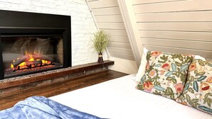 Un dormitorio con chimenea, una cama con almohadas florales y una planta en una mesita de noche.