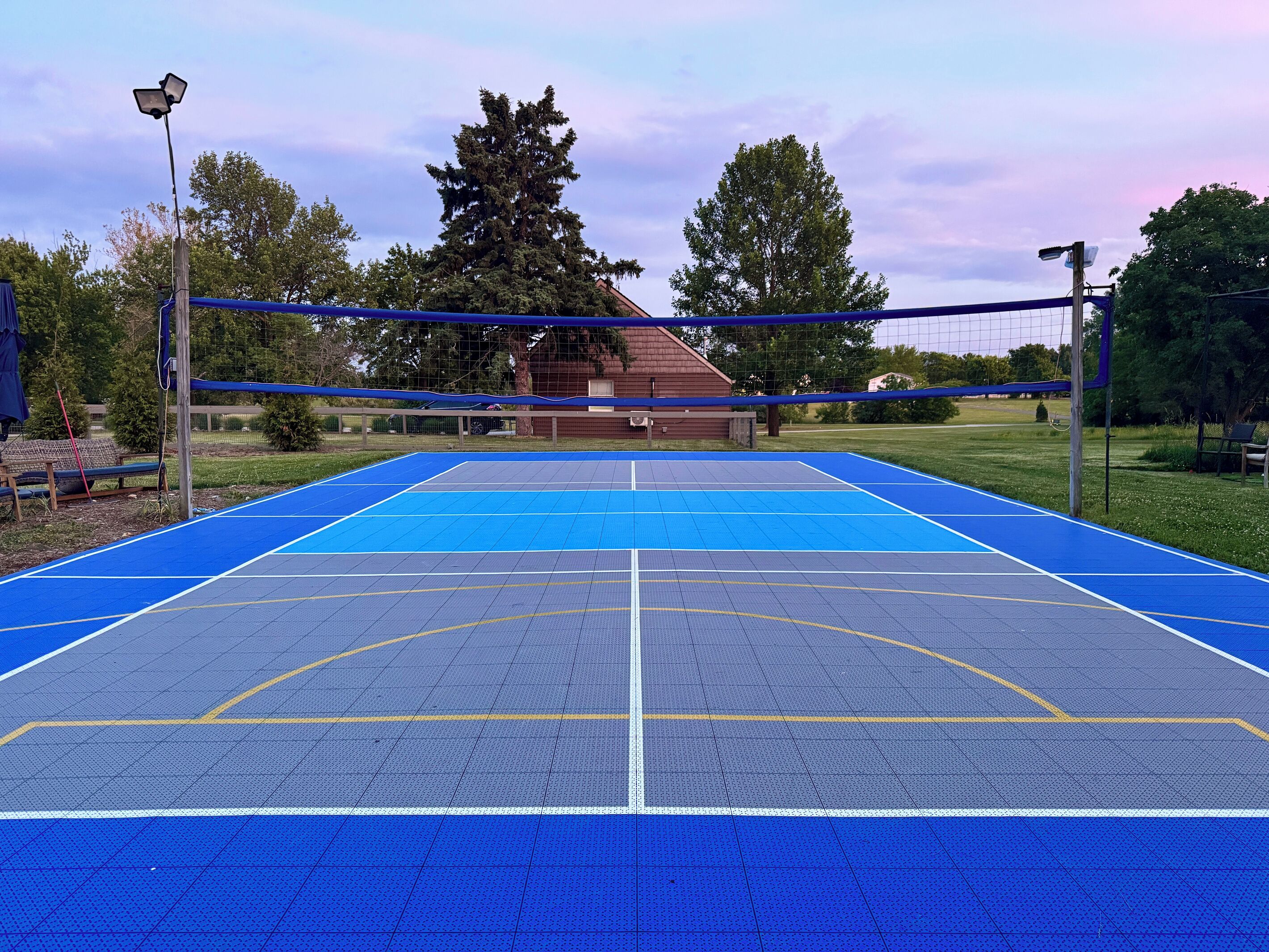 An outdoor tennis court with a net, surrounded by trees and a building in the background.