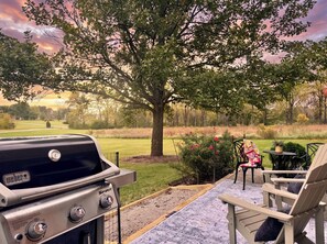 A patio area with a grill, outdoor seating, and a view of a park with trees and a field. - Set on peaceful 5 acres, 5 min. from town, Private Building, Level 2 EV Charger (Bloomington)