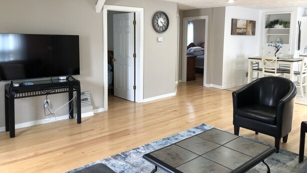 Amplia sala de estar con televisor de pantalla plana, una mesa de centro y un sillón de cuero. Una escalera conduce a un nivel superior, y hay un comedor con mesa y sillas.