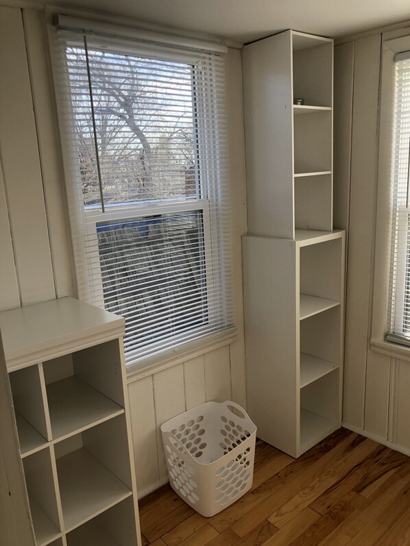 A corner room with built-in shelving, a window with blinds, a wooden floor, and a laundry basket.